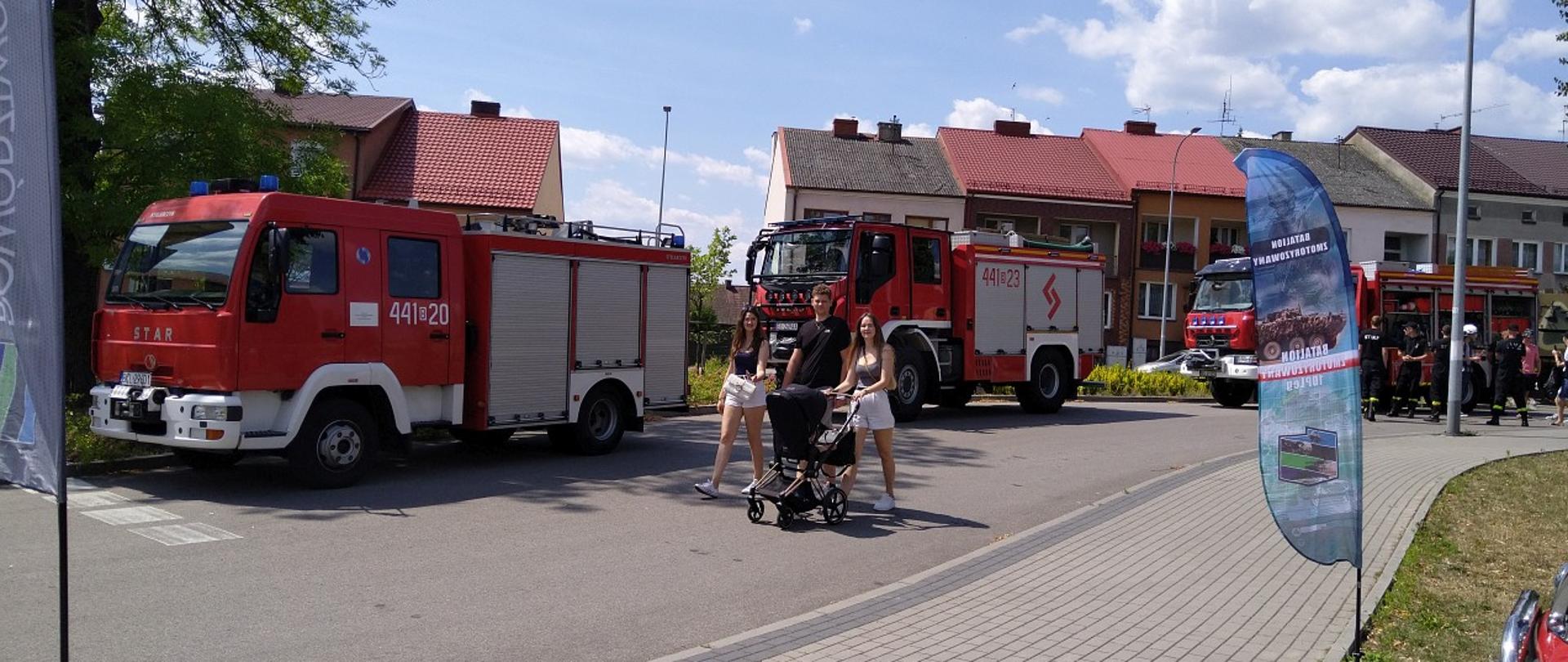 Na zdjęciu widać pojazdy pożarnicze stojące na parku w Kolnie, rodzina z dziećmi spaceruje.
