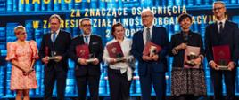 Na tle wielkiego ekranu z napisem Nagrody ministra stoi wiceminister Mrówczyńska i sześć osób trzymających czerwone teczki.