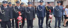 Plac przed pomnikiem Żołnierza Polskiego. Na tle deptaku i kamienic stoją delegacje z wiązankami kwiatów. Strażacy i funkcjonariusze występują w umundurowaniu wyjściowym.