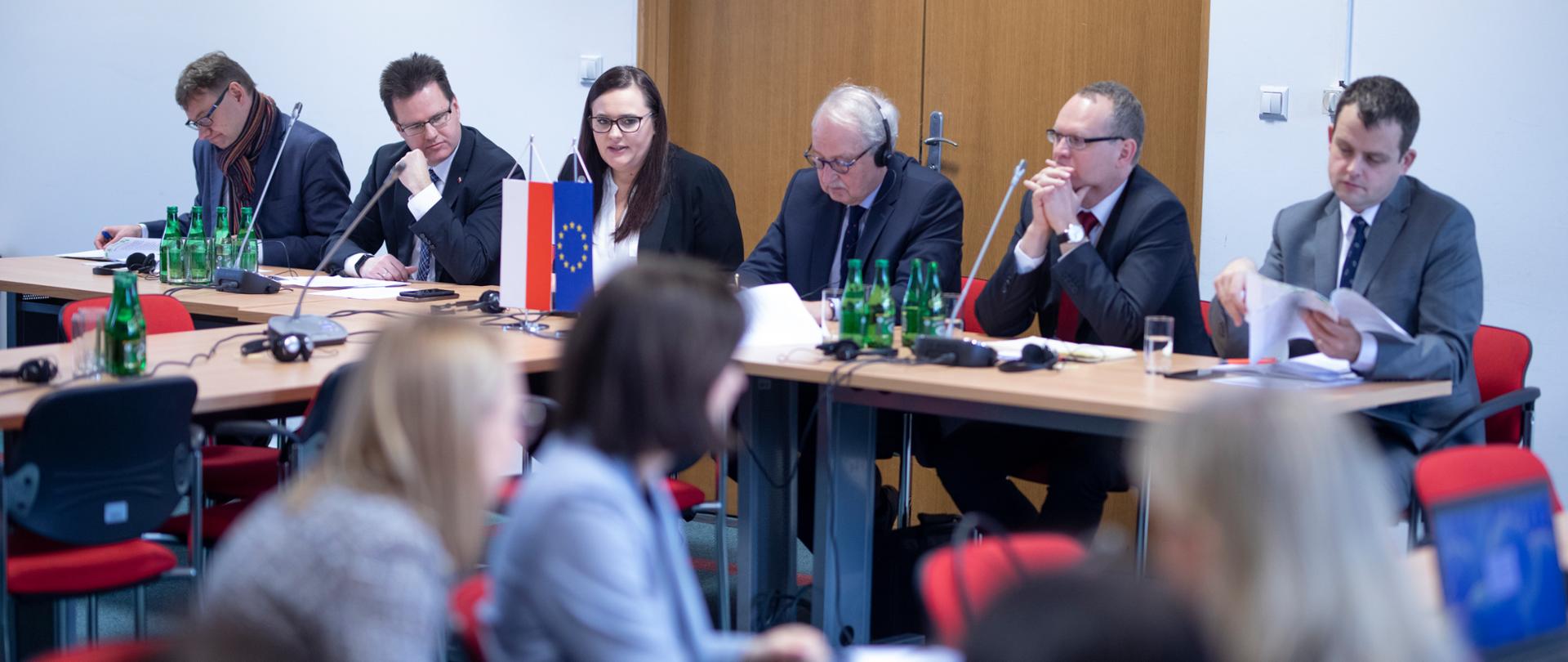 Zdjęcie sali, w której siedzą osoby wśród nich między innymi minister Jarosińska-Jedynak, minister Bittel, dyrektor Orliński i Matthias Ruete. Osoby siedzą przy stole, na którym na proporczykach stoi flaga Polski i Unii Europejskiej. 