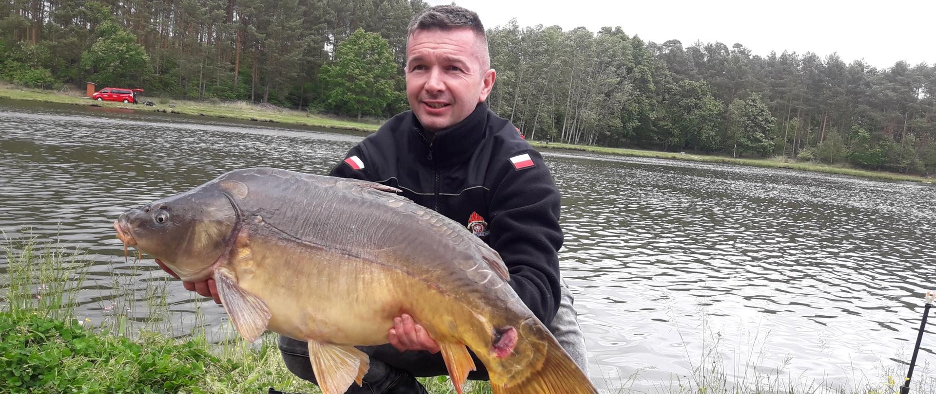 Karp o wadze 13,500 kg złowiony przez zawodnika z KP PSP Wolsztyn. W tle zbiornik wodny położony wśród lasów.