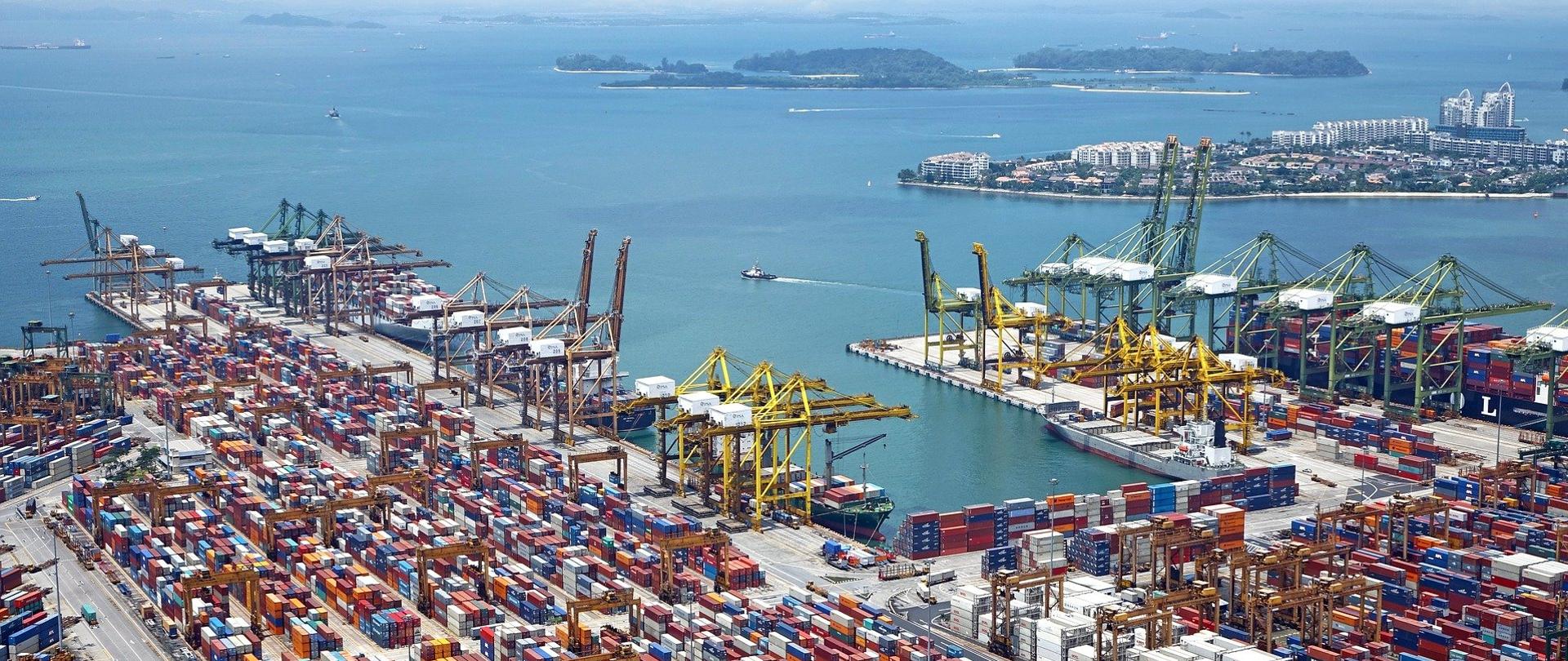 Na zdjęciu znajduje się port - z lewej i prawej strony stoją rzędy kontenerów. W Na drugim planie stoją statki i żurawie portowe. W tle widać małe wyspy porośnięte drzewami.