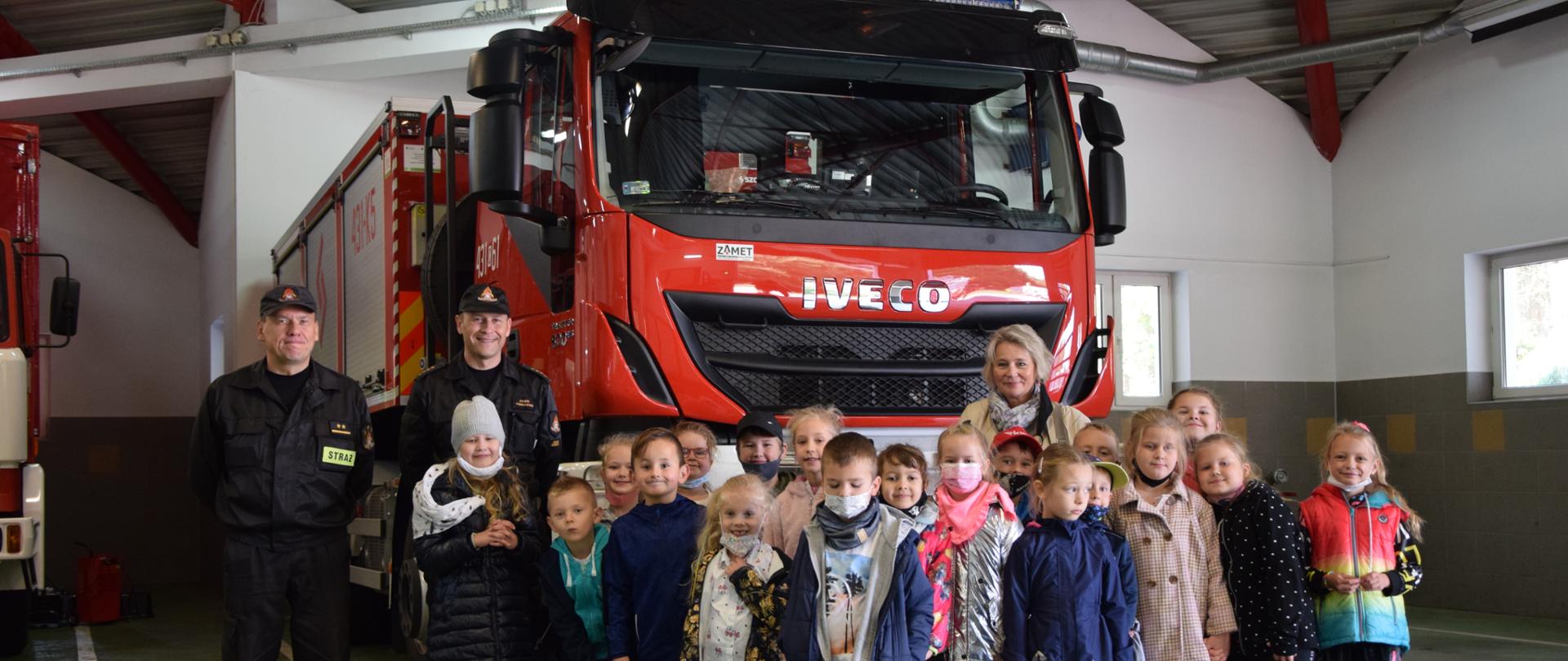 Grupa dzieci wraz z opiekunką oraz dwaj strażacy w ubraniu koszarowym oraz dowódczo-sztabowym stoją na tle wozu strażackiego.