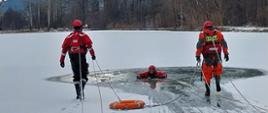 Na zdjęciu widoczni dwaj strażacy na skutym lodzie akwenie wodnym. Obaj w suchych skafandrach koloru pomarańczowego i czerwonego w czerwonych kaskach ochronnych. Obaj przemieszczają się po lodzie z kołem ratunkowym do osoby poszkodowanej w celu ewakuacji osoby z przerębli. 