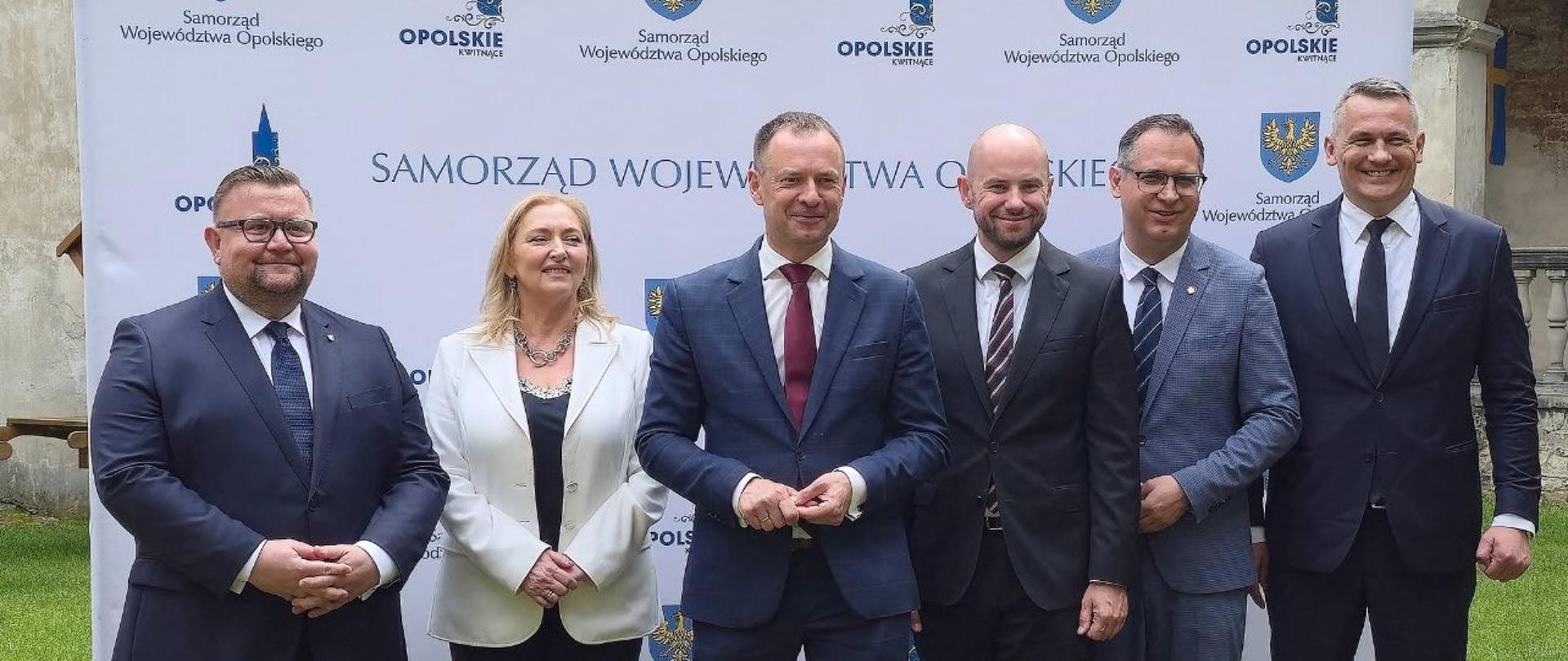 Piotr Borys na konferencji. Na pierwszym planie stoi sześć osób na tle loga województwa opolskiego