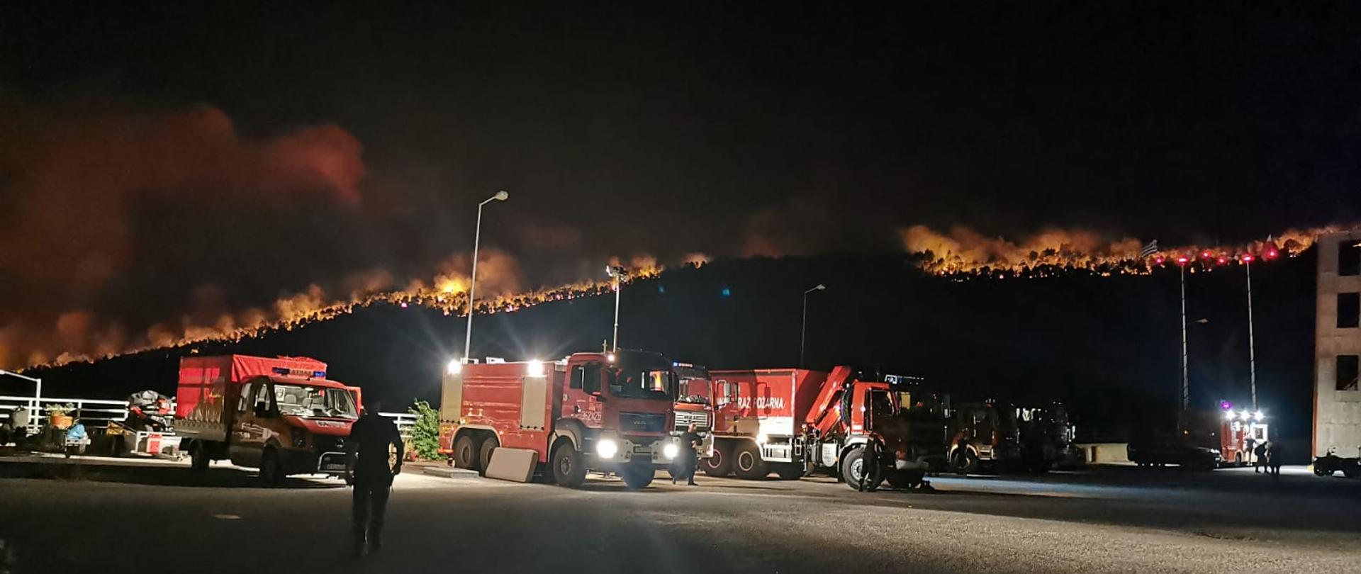 GASZĄ POŻARY W GRECJI. Na zdjęciu widać samochody pożarnicze stojące na placu, za nimi w tle widać płonący las, całe zbocze góry zalesione, bardzo duży pożar - płomienie, dym. 