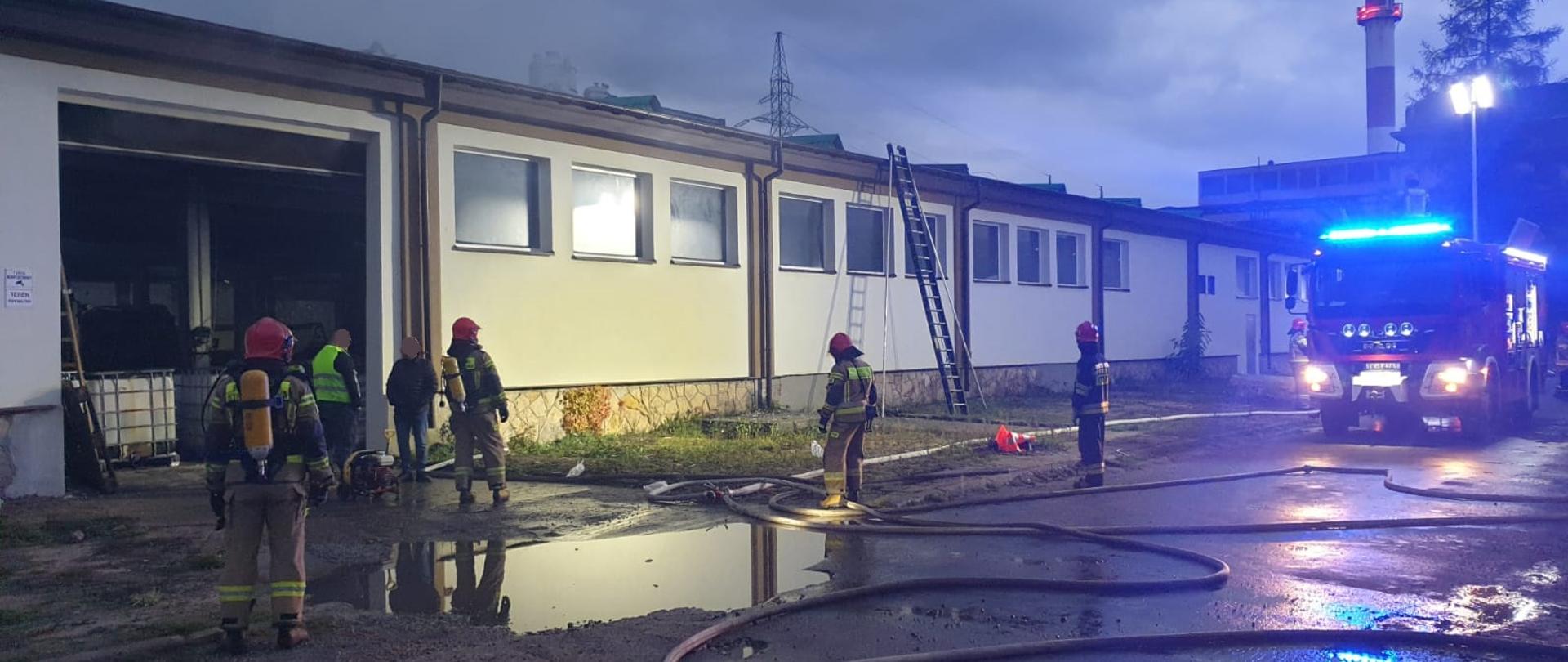 Zdjęcie przedstawia strażaków przed wejściem do hali produkcyjno-magazynowej. Przed nimi są rozwinięte linie gaśnicze i kałuża wody. Obok nich stoi samochód pożarniczy oświetlający teren akcji.