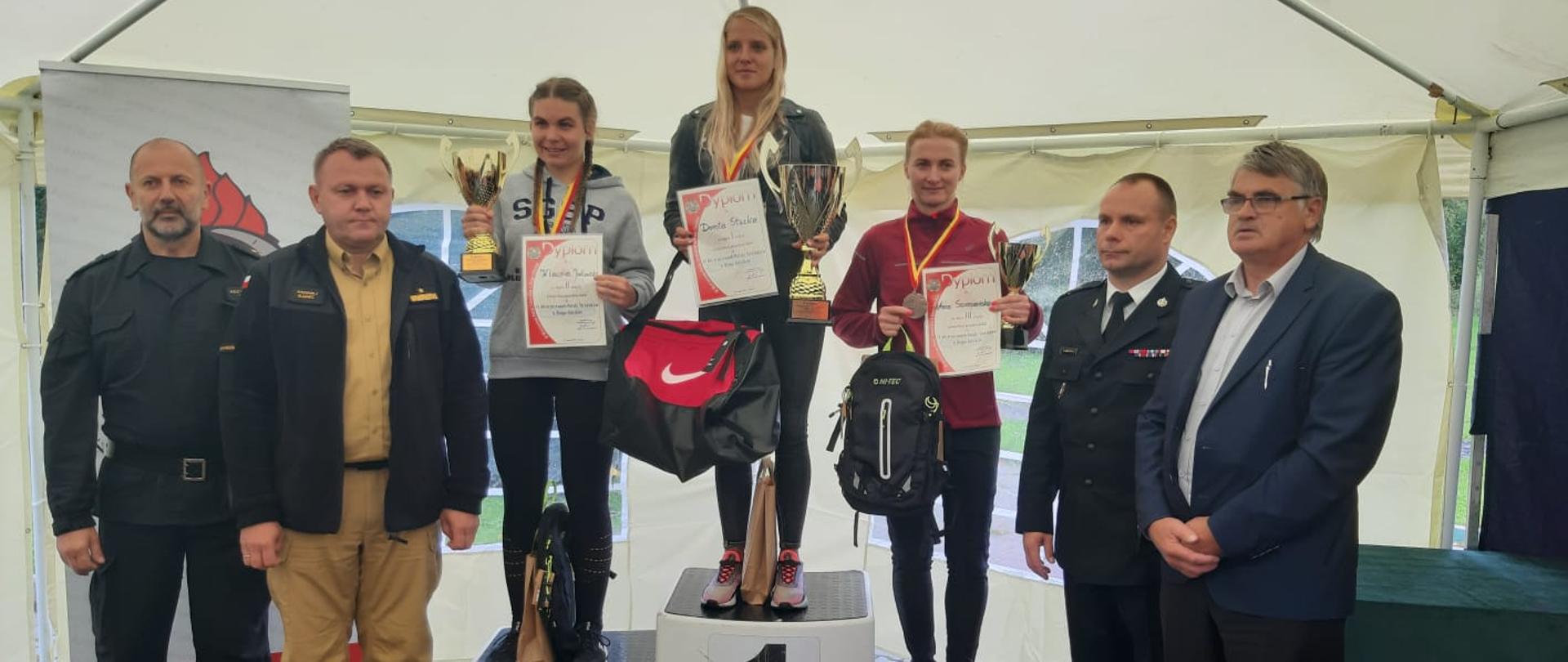Zdjęcie przedstawia podium znajdujące się pod białym zadaszeniem. Na 3 stopniach stoją kobiety w sportowych strojach z pucharami i dyplomami w rękach. Obok stoi czterech mężczyzn – 3 w mundurach i 1 garniturze. 