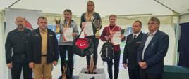 Zdjęcie przedstawia podium znajdujące się pod białym zadaszeniem. Na 3 stopniach stoją kobiety w sportowych strojach z pucharami i dyplomami w rękach. Obok stoi czterech mężczyzn – 3 w mundurach i 1 garniturze. 