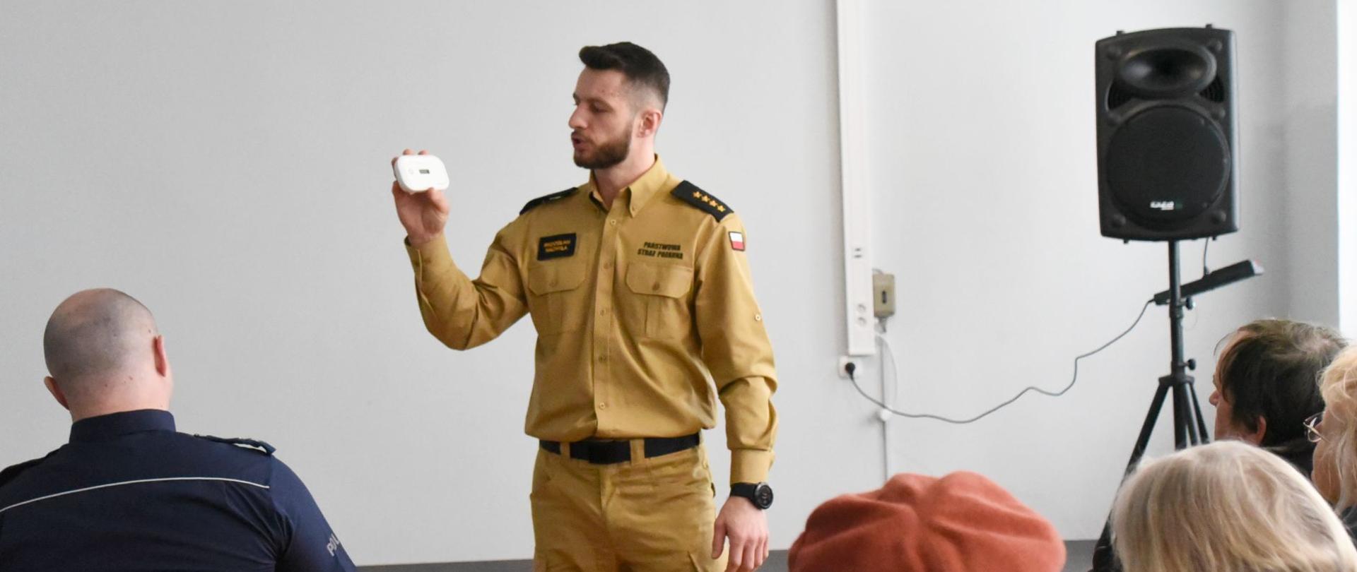 Zdjęcie wykonane podczas prelekcji realizowanych w ramach akcji babciu, dziadku nie czujesz - trujący, bezwonny, bezbarwny. Na zdjęciu widać funkcjonariusza z komendy miejskiej Państwowej Straży Pożarnej w Radomiu. Mężczyzna ubrany jest w ubranie dowódcze w kolorze żółtym piaskowym z czarnymi emblematami. Funkcjonariusz w jednej ręce trzy czujkę tlenku węgla oraz opowiada o zagrożeniach związanych z tlenkiem węgla. Przed nim widać siedzących na sali słuchaczy – uczestników spotkania. Są to przedstawiciele policji oraz osoby starsze. 