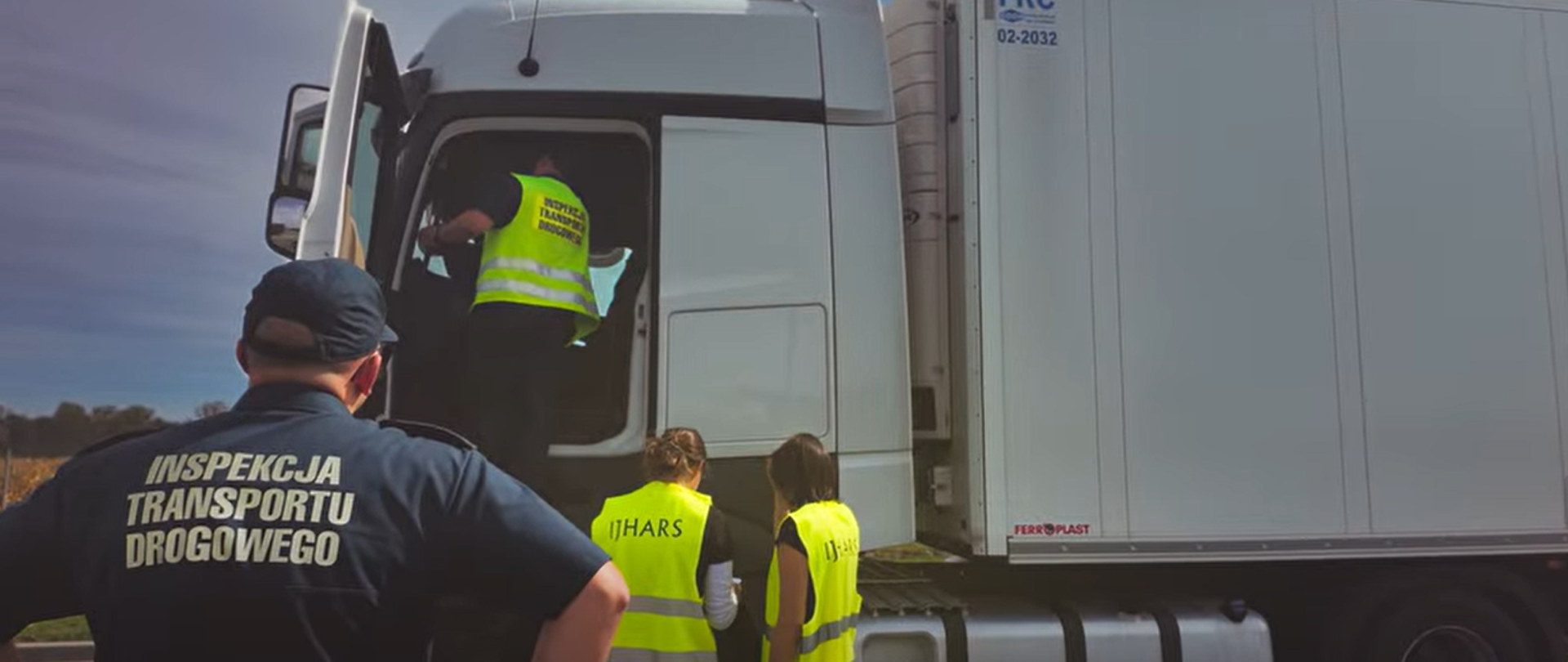 Inspektor Transportu Drogowego oraz pracownicy IJHARS podczas wspólnej kontroli białego samochodu ciężarowego. Jeden z inspektorów ITD sprawdza kabinę pojazdu, a przy samochodzie stoją dwie osoby w kamizelkach odblaskowych IJHARS