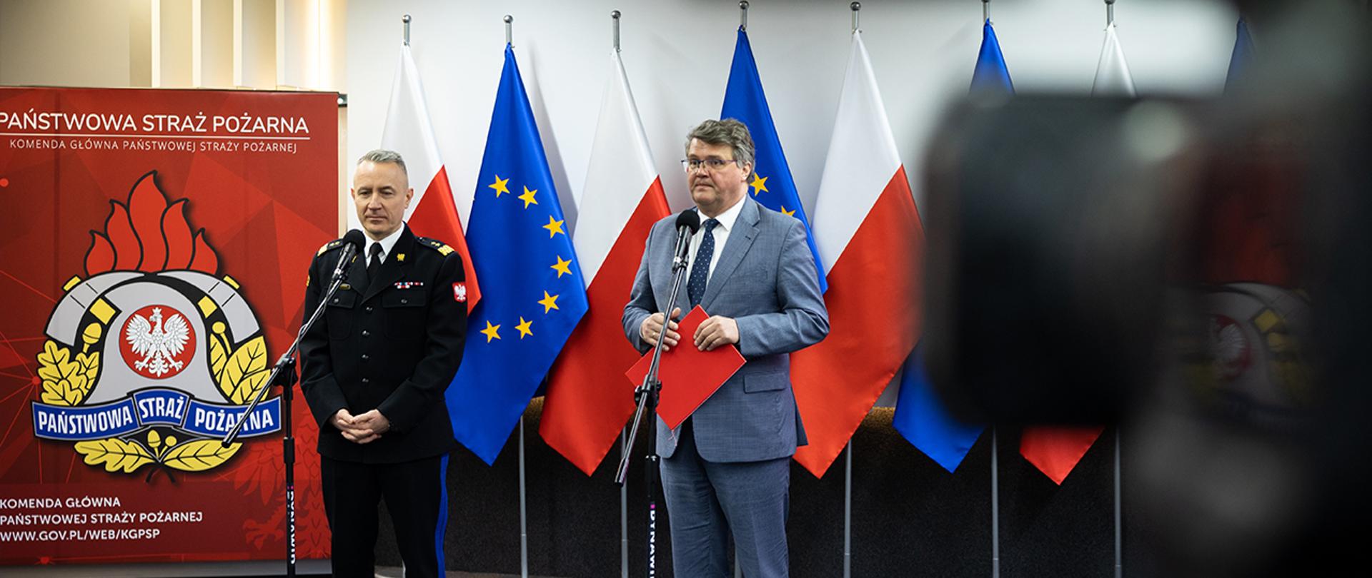 Wiceminister Maciej Wąsik oraz komendant główny PSP na tle flag Polski i UE podczas konferencji prasowej.