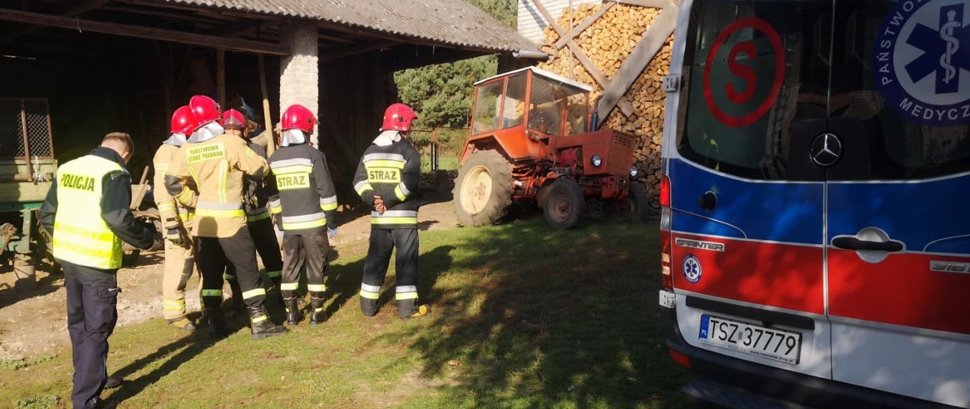 Zdjęcie przedstawia podwórko gdzie doszło do tragedii. Z lewej strony widać grupę strażaków, w głębi stoi ciągnik rolniczy, a po prawej stronie widać ambulans Państwowego Ratownictwa Medycznego 