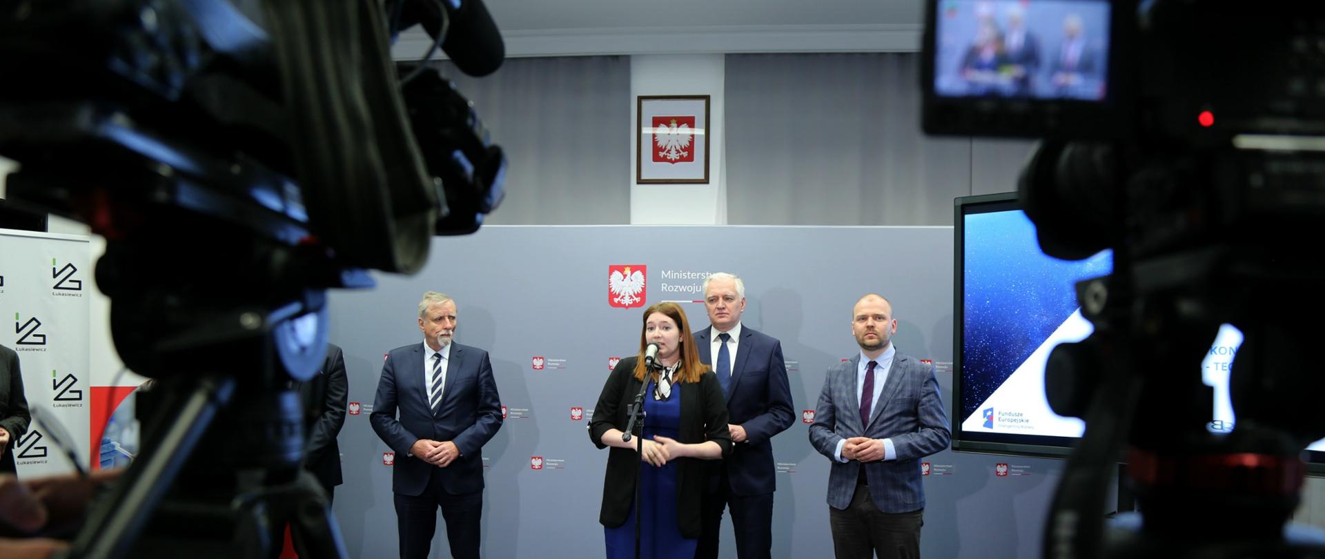 Minister Gembicka mówi do mikrofonu, przed nią stoją kamery, za nią stoi uczestnicy spotkania wśród nich między innymi premier Gowin