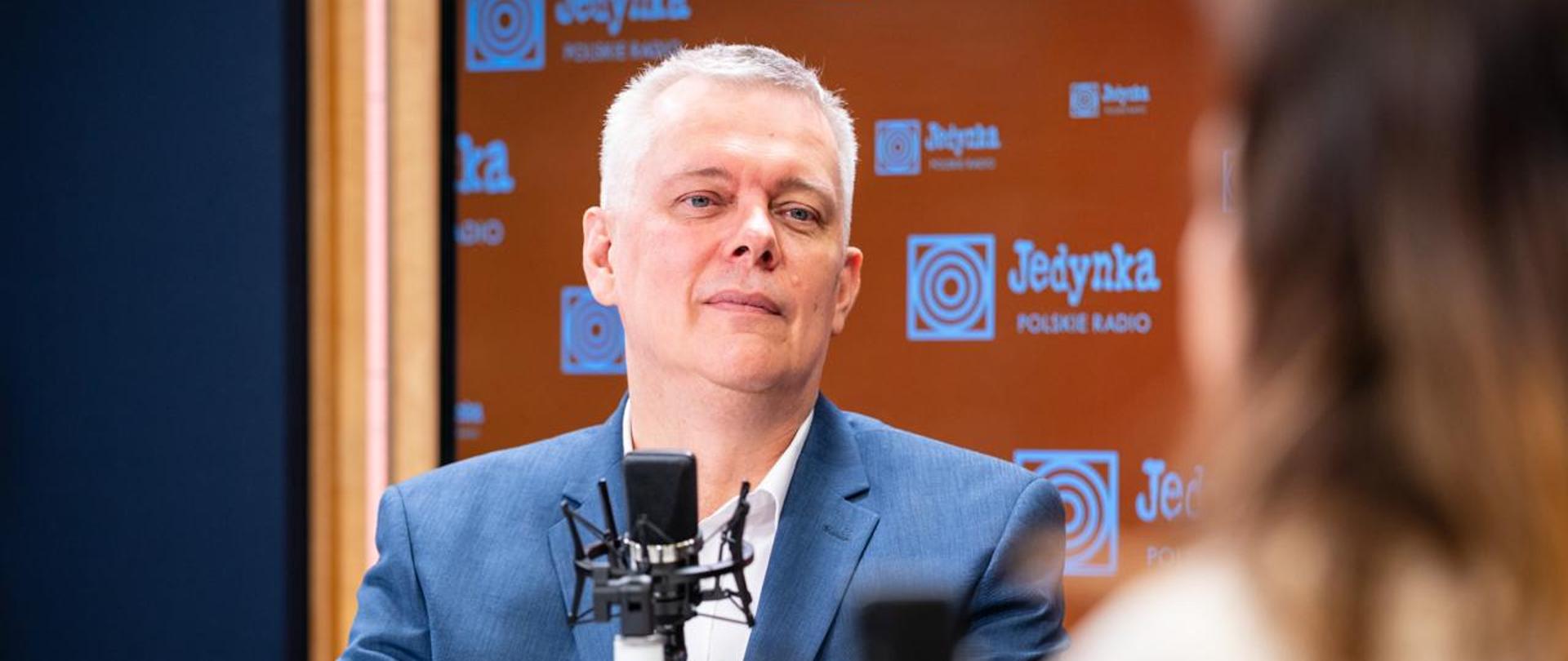 Minister Tomasz Siemoniak siedzi przed czarnym mikrofonem w studiu Programu 1 Polskiego Radia. W tle za ministrem - po prawej stronie pomarańczowa ścianka z logo i napisem Jedynka Polskie Radio (zmultiplikowane), a po lewej stronie granatowa ściana. Po prawej stronie zdjęcia widać siedzącą tyłem redaktor Agatę Kondzińską. W lewym dolnym rogu widać pomarańczowy kubek Jedynki Polskiego Radia.