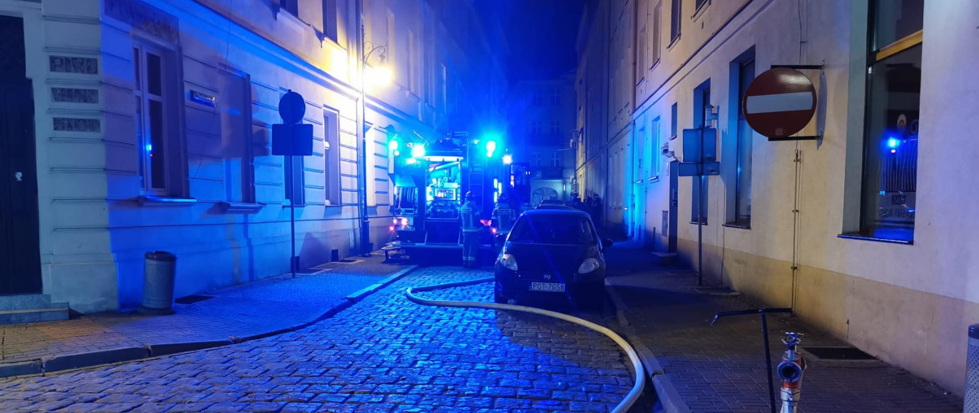 Na zdjęciu widzimy tył samochodu pożarniczego stojącego na brukowanej ulicy pomiędzy kamienicami. Do samochodu dochodzi także wąż strażacki koloru białego, którego druga końcówka podłączona jest do hydrantu. Na zdjęciu widzimy także zaparkowany samochód osobowy koloru czarnego. Z samochodu strażackiego błyskają niebieskie światła ostrzegawcze. 