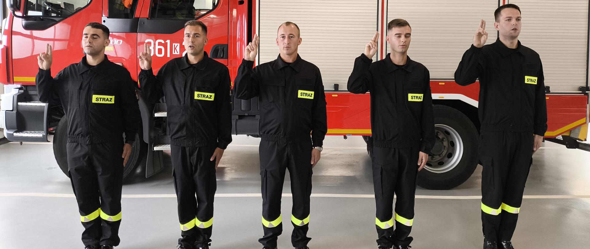W szeregu stoją strażacy ubrani w czarne ubrania koszarowe, prawe ręce mają uniesione do ślubowania. Z tyłu widoczny czerwony samochód pożarniczy.