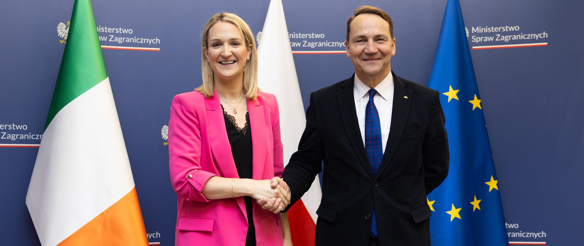 In the photo, there is a woman in a pink blazer and a man in a black suit. They are looking toward the camera and shaking hands, Behind them are the flags of Irleand, Poland, and the EU. 
