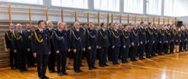 Strażacy Państwowej Straży Pożarnej oraz ochotniczych straży pożarnych stoją w czterech rzędach podczas uroczystości. Uroczystość na sali gimnastycznej Komendy Wojewódzkiej Państwowej Straży Pożarnej w Opolu.