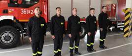 W szeregu stoją strażacy ubrani w czarne ubrania koszarowe. Z tyłu widoczny czerwony samochód pożarniczy.