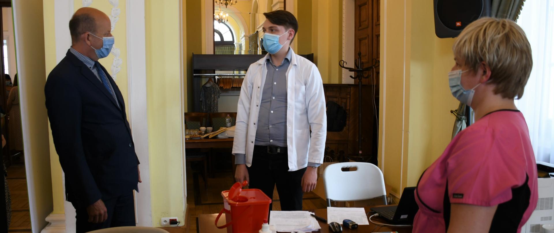 Od lewej strony: Wojewoda Mazowiecki rozmawiający z lekarzem, przed mężczyznami stoi biurko na którym leżą niezbędne rzeczy do wykonania szczepienia. Po prawej stronie w rogu pielęgniarka w różowym fartuchu. 