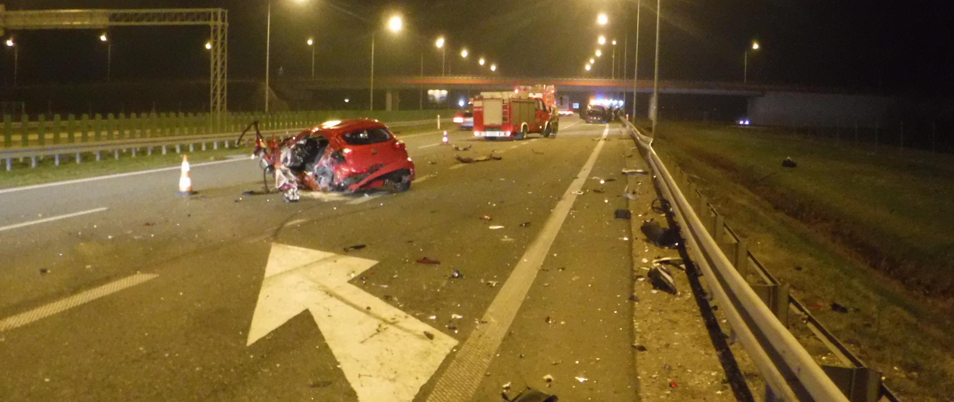 Na zdjęciu widać rozbity czerwony samochód osobowy stojący na autostradzie. Na jezdni znajdują się rozbite elementy karoserii. W tle widać dwa czerwone samochody strażackie, drugi rozbity samochód oraz wiadukt nad autostradą. Jest ciemno, ale jezdnia jest oświetlona przez latarnie.