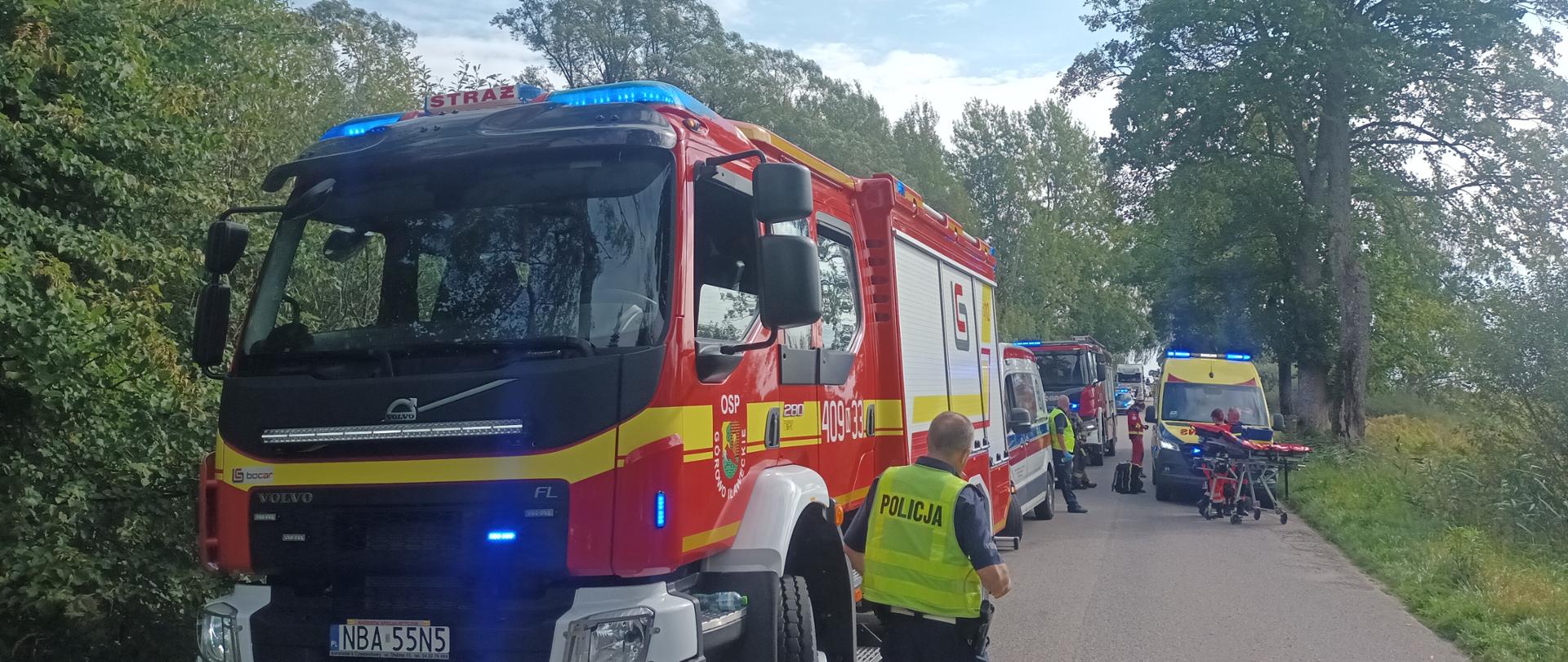 Na zdjęciu widoczna jest akcja ratunkowa na wiejskiej drodze otoczonej drzewami. Na pierwszym planie stoi wóz strażacki OSP z włączonymi sygnałami świetlnymi. Obok pojazdu znajduje się funkcjonariusz policji w kamizelce odblaskowej, kierujący się w stronę miejsca zdarzenia. W głębi kadru widać kolejne pojazdy ratownicze: karetki pogotowia i drugi wóz strażacki. Na poboczu i jezdni rozstawione są nosze oraz sprzęt medyczny. Scena przedstawia działania służb ratunkowych – straży pożarnej, policji i ratownictwa medycznego – po zdarzeniu drogowym.