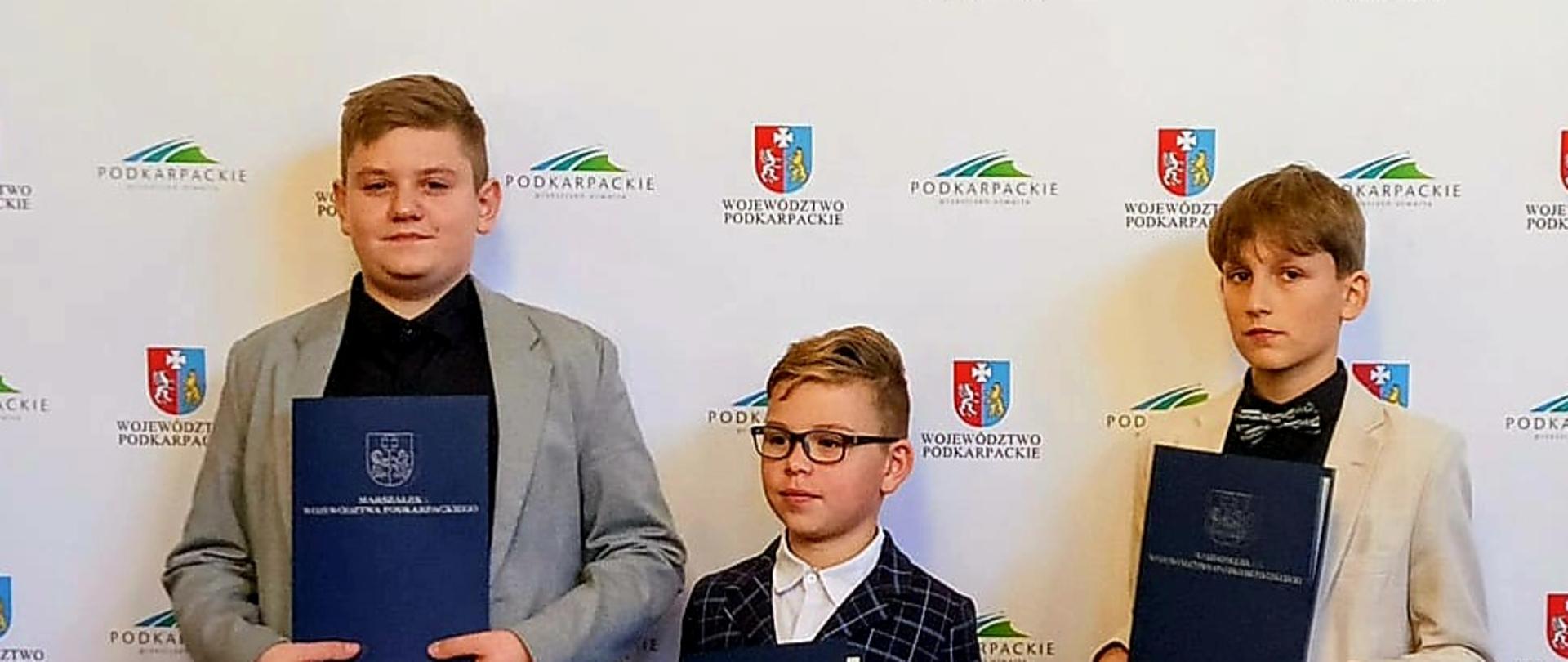 Trzech młodych stypendystów (dwóch chłopców, nastolatków w garniturach i jeden młodszy chłopiec w okularach i kratkowanym garniturze) stojących obok siebie, trzymających w dłoniach dyplomy. W tle ściana z logo Województwa Podkarpackiego.
