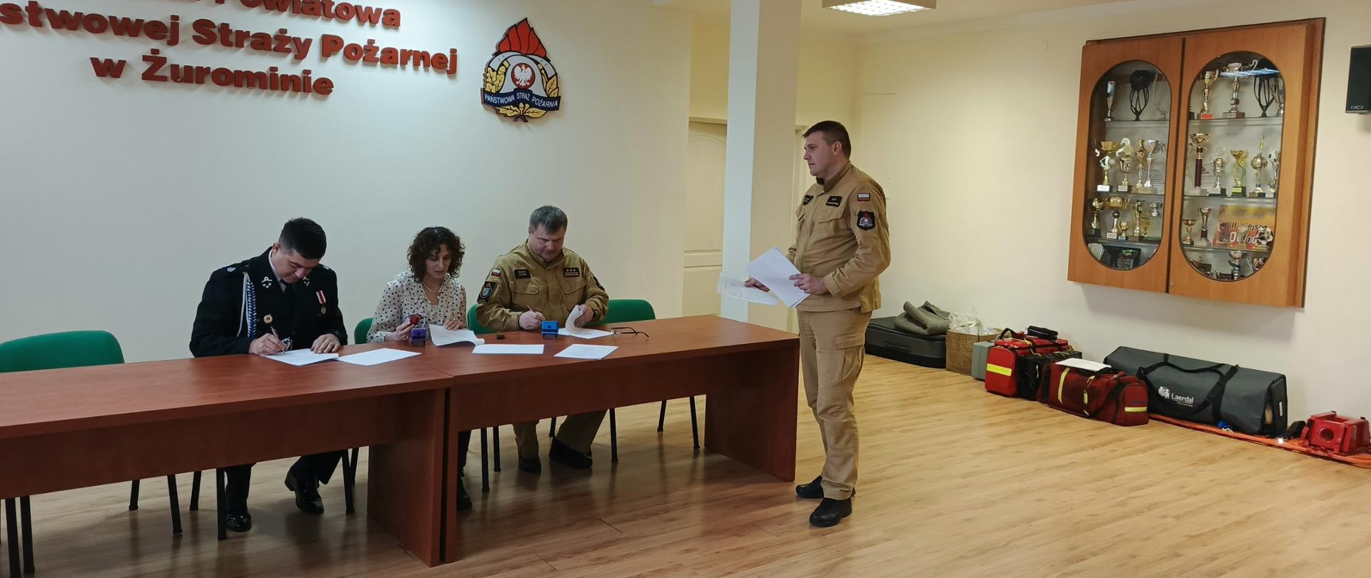 Zdjęcie przedstawia oficjalne podpisanie porozumienia w Komendzie Powiatowej Państwowej Straży Pożarnej w Żurominie, w którym uczestniczą przedstawiciele straży oraz osoba cywilna. Na pierwszym planie widać osoby siedzące przy stole i podpisujące dokumenty, a w tle znajdują się gabloty z pucharami oraz sprzęt ratowniczy.