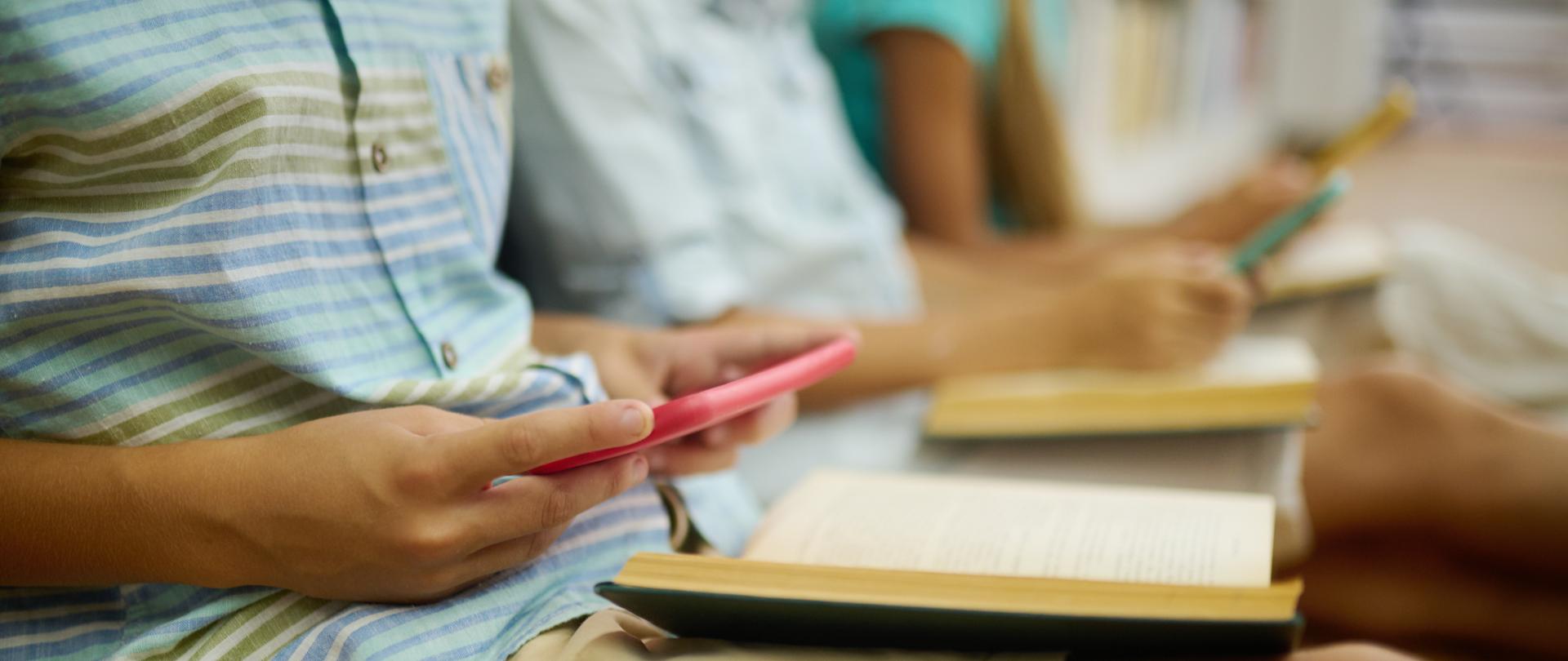 Zdjęcie ilustracyjne w poziomie. Dzieci siedzą przy biblioteczce. W ręku trzymają smartfony.
