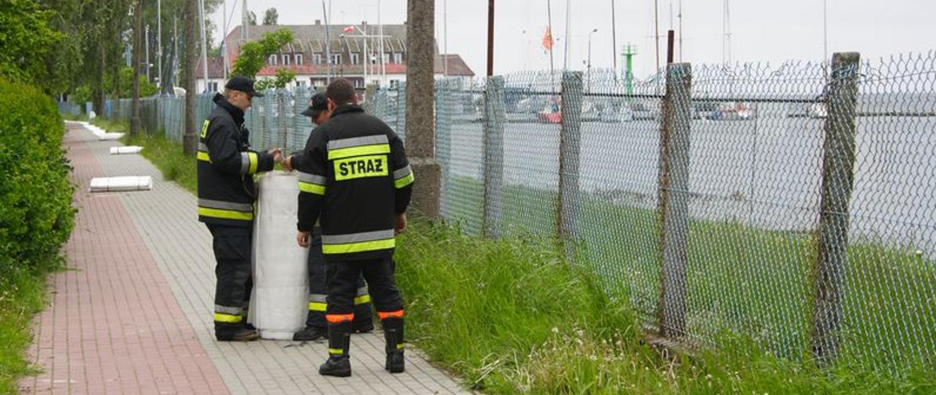 Zdjęcie przedstawia strażaków ustawiających zaporę przeciwpowodziową napełnianą wodą