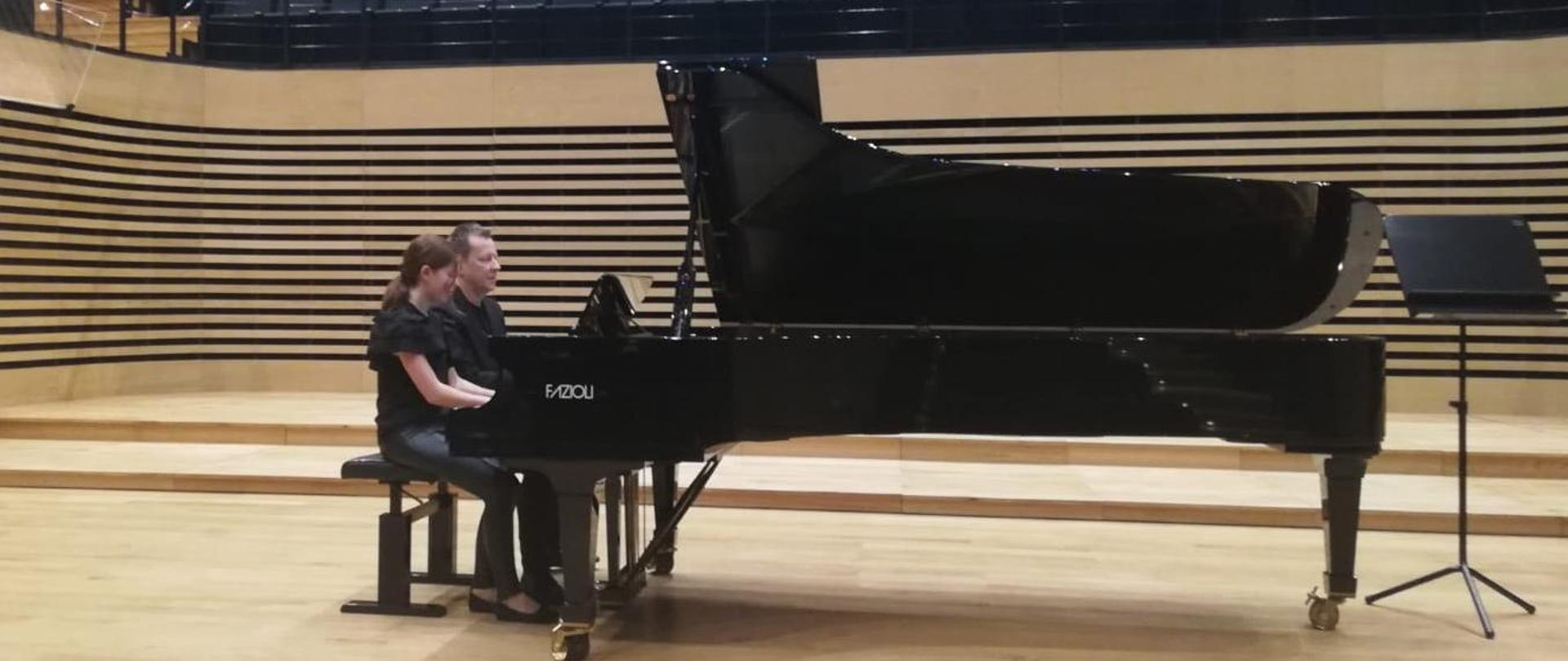 Uczennica gra na fortepianie razem z nauczycielem na cztery ręce na scenie sali koncertowej PSM