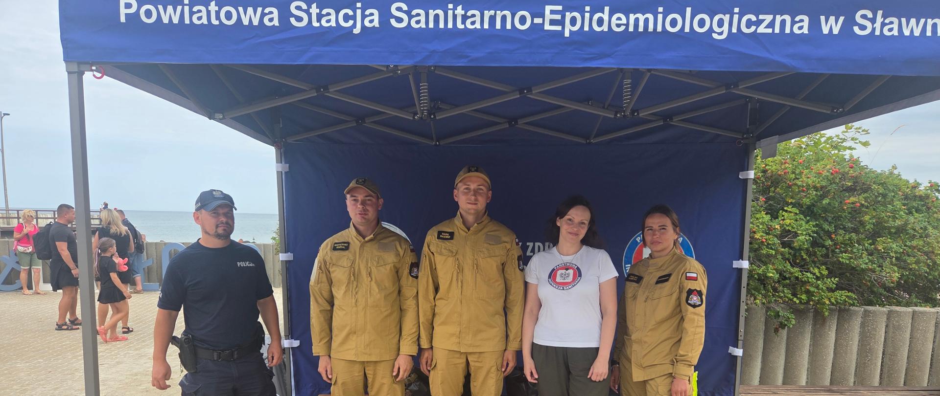 Przedstawiciele Państwowej Straży Pożarnej oraz Powiatowej Stacji Sanitarno-Epidemiologicznej w Sławnie podczas akcji „Bezpieczne wakacje" w Darłówku. Uczestnicy stoją przed namiotem informacyjnym.