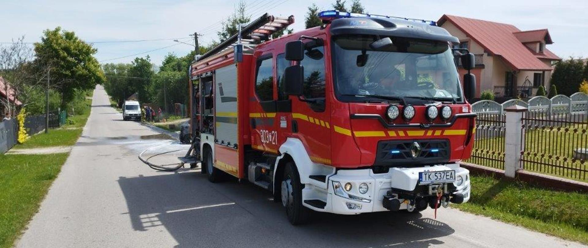 Zdjęcie przedstawia ulicę na której zaparkował samochód gaśniczy. Zza niego wystaje fragment spalonego samochodu osobowego.