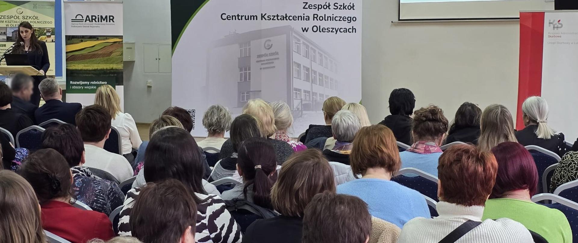 Uczestnicy szkolenia siedzą w sali. Przed nimi przy mównicy stoi ekspertka. Na ekranie wyświetlana jest prezentacja.