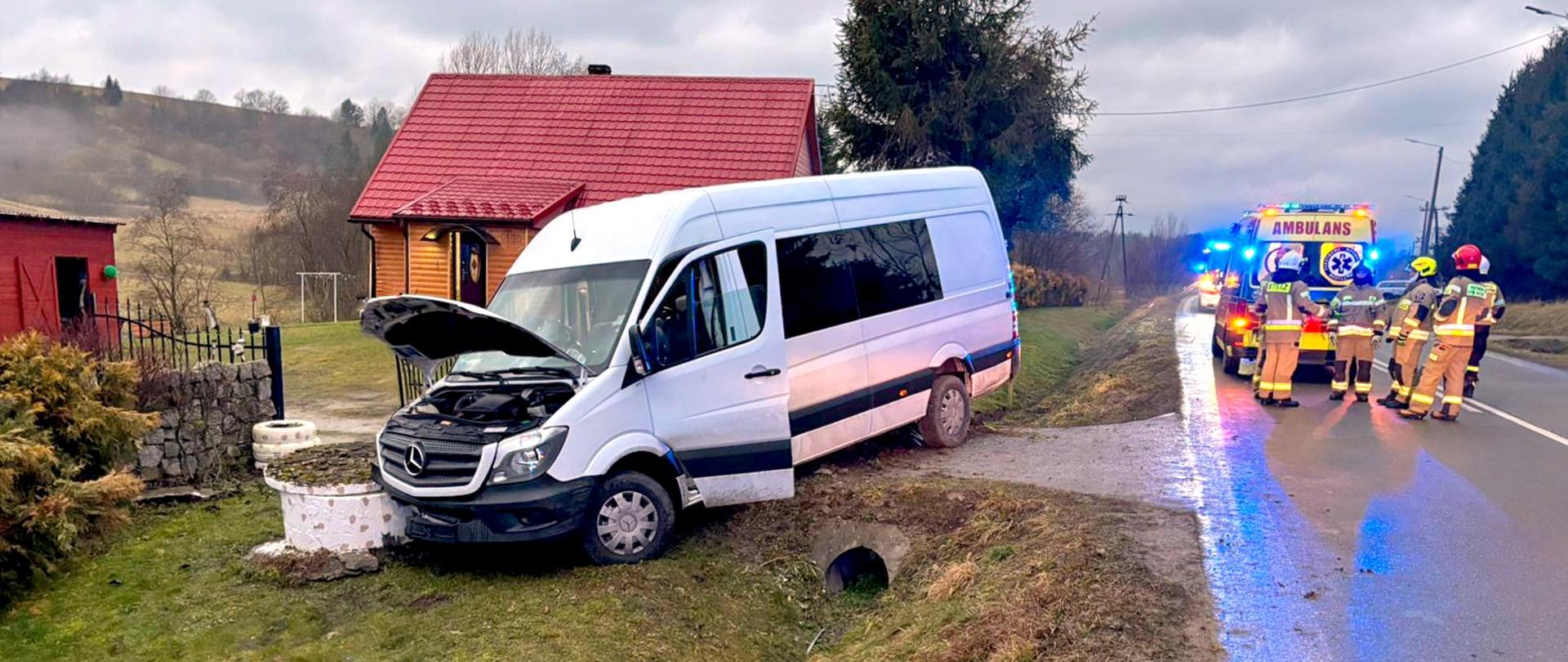 Na zdjęciu znajduje się bus przeznaczony do przewozu osób poza drogą. Uderzył w studnię i ma podniesioną maskę. Za busem w tle widać dom. Z tyłu na drodze po prawej stronie stoją strażacy i karetka pogotowia. 