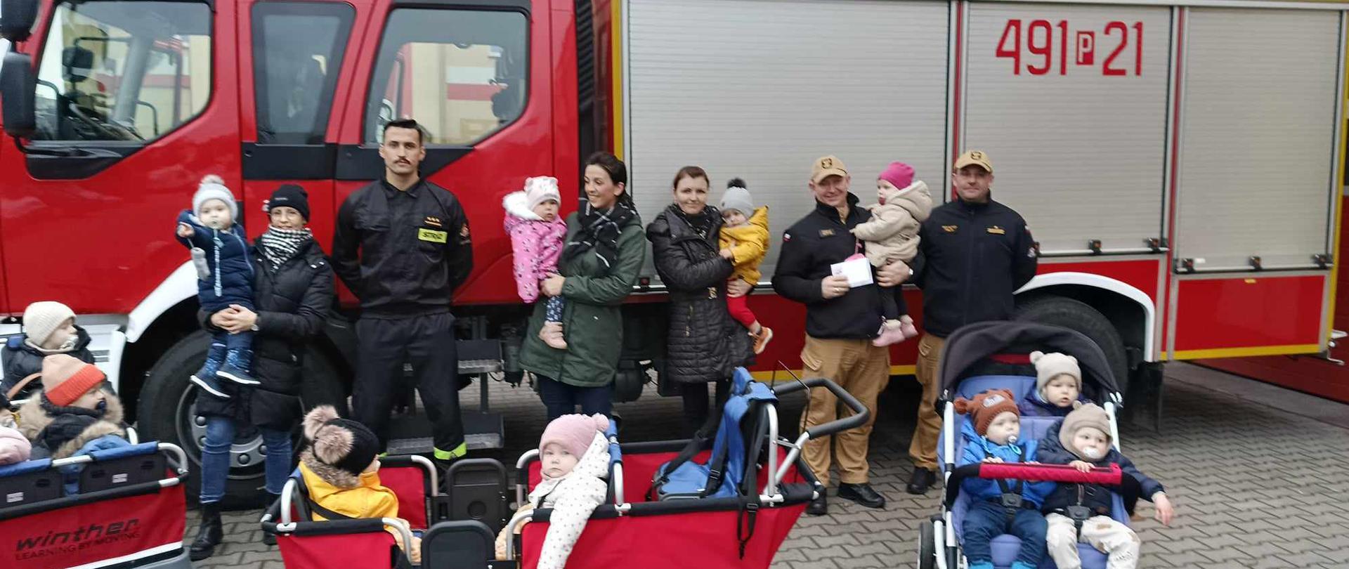 Zdjęcie przedstawia dzieci wraz z opiekunami i strażakami na tle pojazdu pożarniczego