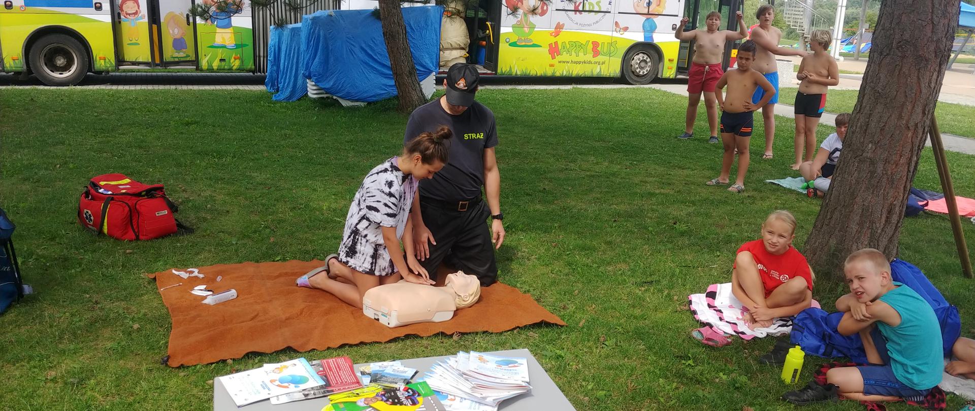 Plac przy basenie miejskim, aspirant Mateusz Rączka udziela zgromadzonym dzieciom instruktażu jak udzielać pierwszej pomocy poszkodowanym, na stoliku ulotki dla dzieci z akcji Kręci mnie bezpieczeństwo. 