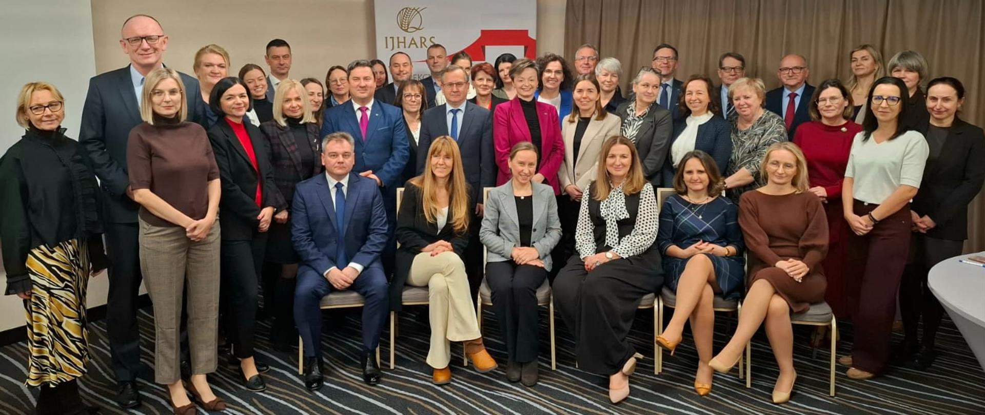 Grupowe zdjęcie elegancko ubranych ludzi. Stoją lub siedzą w kilku rzędach. Wszyscy patrza w obiektyw