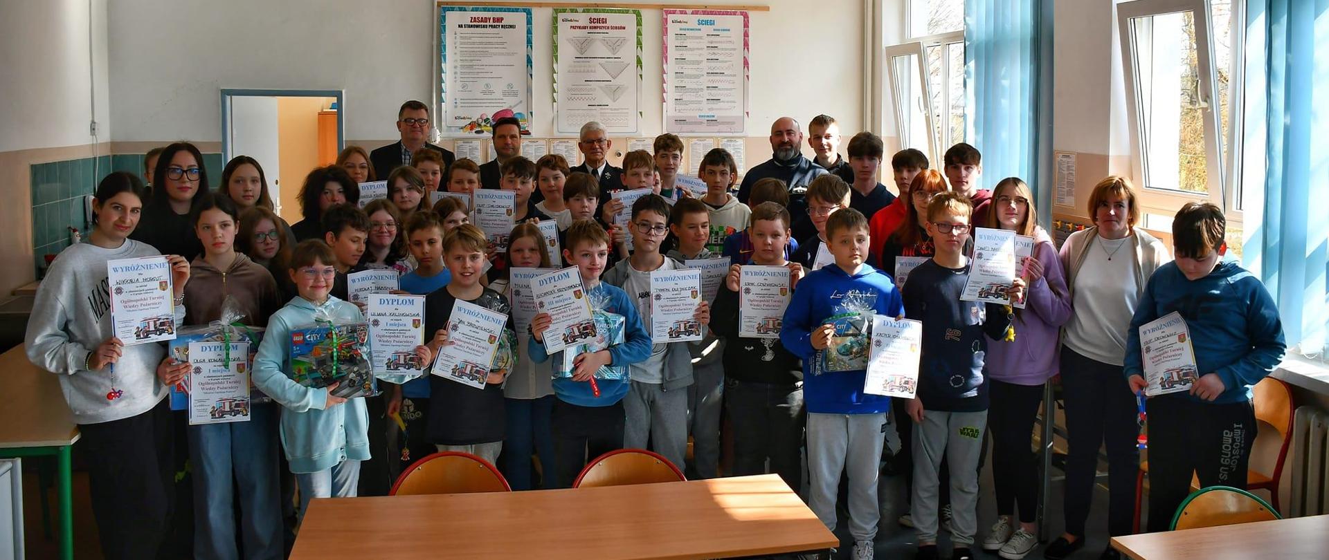 Zdjęcie zrobione w sali szkolnej. Na zdjęciu duża grupa dzieci z dyplomami w rekach. Za nimi organizatorzy konkursu.