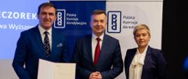 Na tle baneru z napisami Polska Komisja Akredytacyjna stoi minister Wieczorek pomiędzy mężczyzną w granatowym garniturze i wiceminister Mrówczyńską.