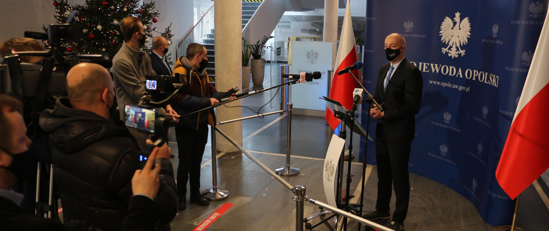 Konferencja prasowa wojewody Adriana Czubaka. Na zdjęciu po prawej stronie stojący na granatowej ściance wojewoda, przemawiający do dziennikarzy. przed nim stoi mikrofon na statywie oraz barierki, odgradzające. Po bokach ścianki stoją biało-czerwone flagi. Na przeciw wojewody dziennikarze (kadr boczny). W tle widać choinkę oraz dalszą część budynku urzędu. 