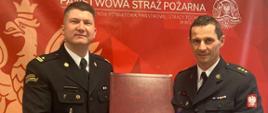 Zdjęcie wykonane wewnątrz pomieszczenia. Przedstawia Z-ce komendanta Powiatowego PSP w Wolsztynie wraz z Komendantem Powiatowym PSP w Wolsztynie. W tle baner w kolorze czerwonym z napisem Państwowa Straż Pożarna.