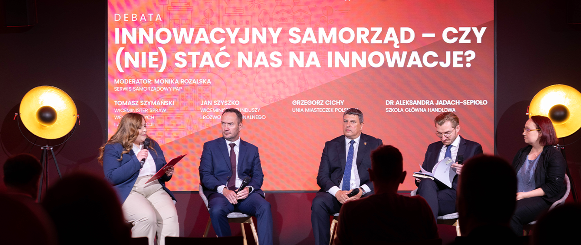 Grupa osób siedzi na scenie. Wśród nich jest wiceminister Tomasz Szymański. W tle czerwony ekran z napisem Innowacyjny samorząd – czy (nie) stać nas na innowacje? - debata