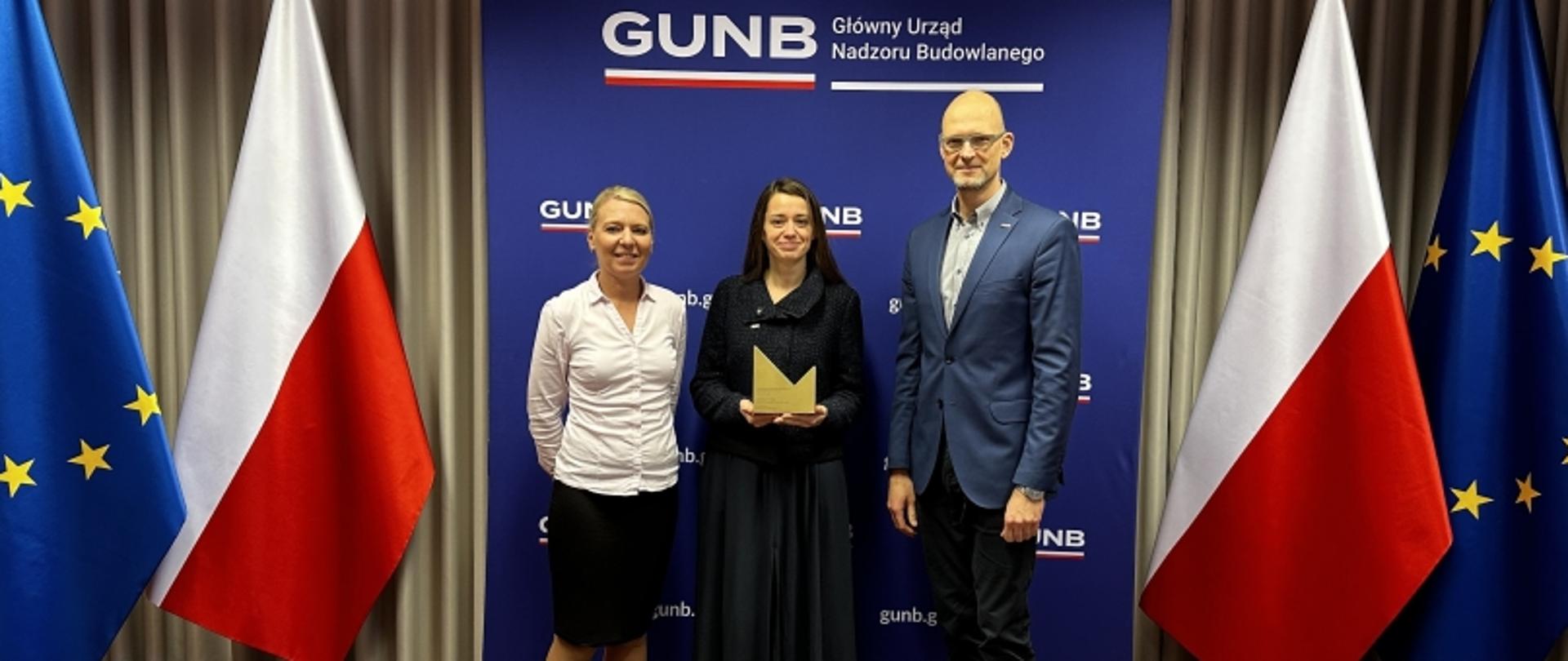 Laureaci na tle ścianki GUNB z flagami Polski i UE; osoba w środku ze statuetką.