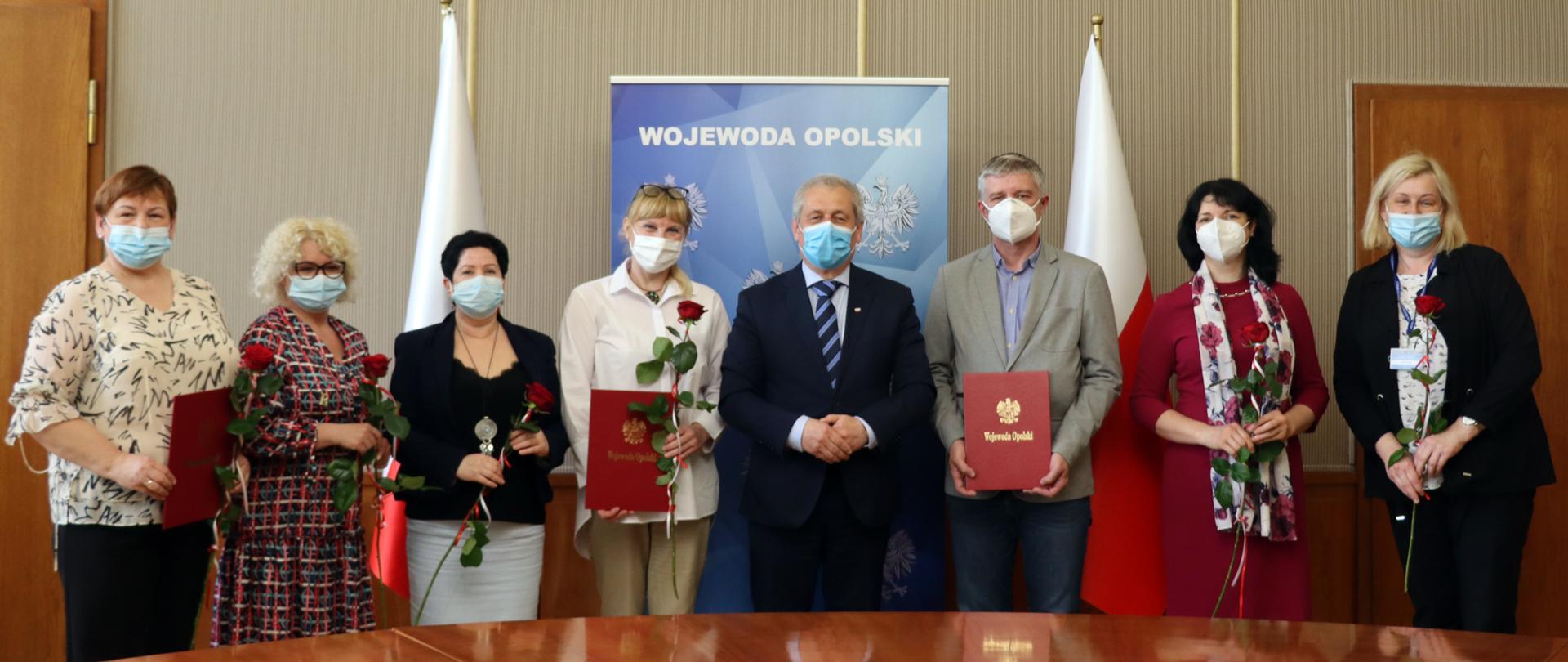 Na zdjęciu 8 osób, stojących na tle ścianki Wojewoda Opolski w Sali im. Braci Kowalczyków w OUW. W centralnej części kadru dwóch mężczyzn, jest to wojewoda opolski oraz jeden z konsultantów. Od lewej strony kadru, w szeregu stoją pielęgniarki, każda w dłoni trzyma różę, jaką otrzymały od wojewody. W tle biało-czerwone flagi. 