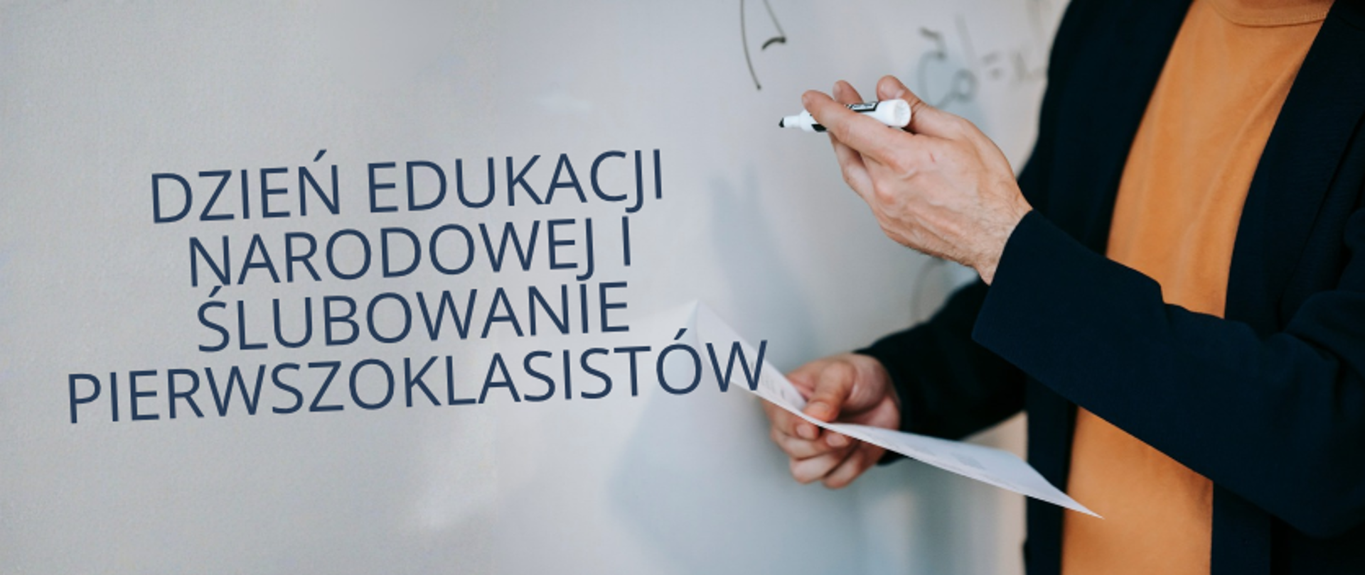 Baner z napisem „Dzień Edukacji Narodowej. Ślubowanie Pierwszoklasistów”. Po prawej stronie widoczna osoba ubrana w czarną marynarkę i pomarańczową bluzkę, trzymająca marker oraz kartkę papieru. W tle znajduje się jasna tablica.