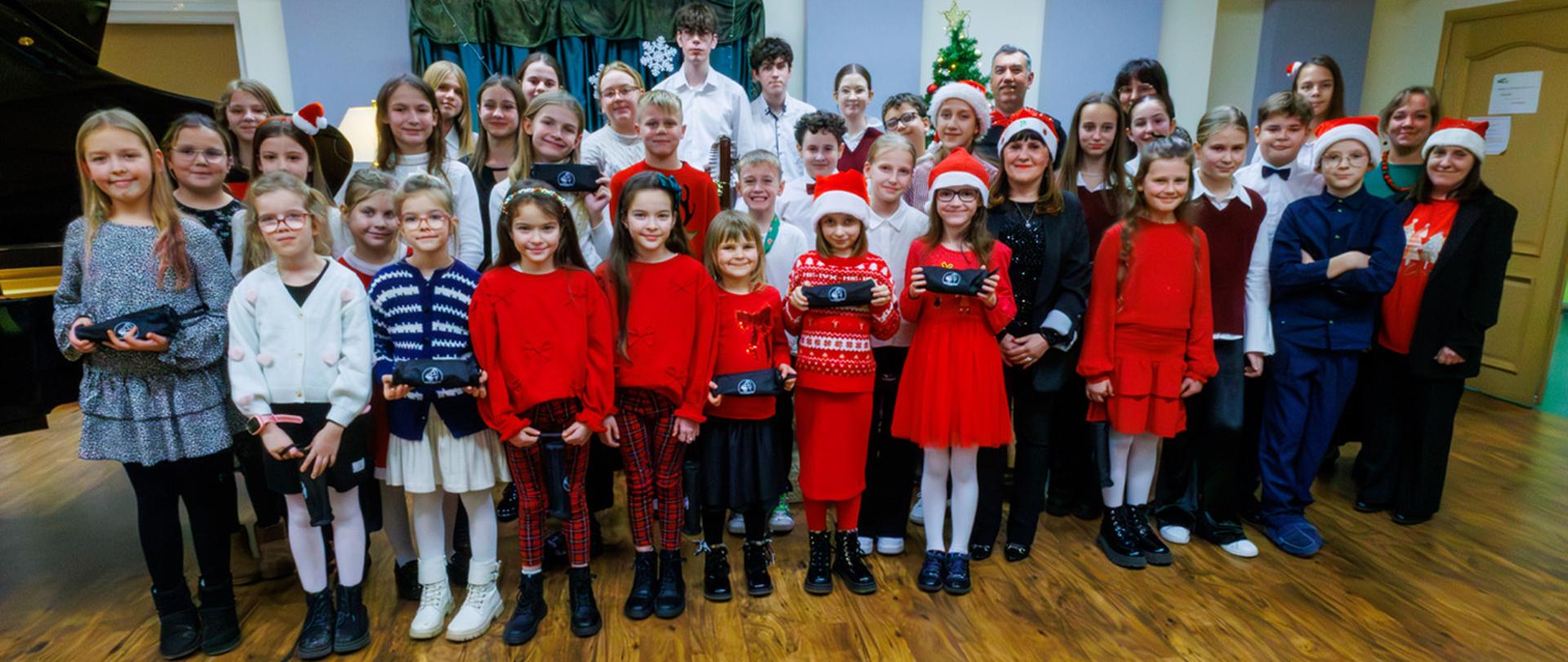 Grupa kilkudziesięciu osób, uczniów i nauczycieli stoi w auli szkolnej. W tle dekoracje świąteczne, część dzieci ubrana w stroje bożonarodzeniowe.