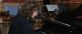 Zdjęcie przedstawia mężczyznę grającego na czarnym fortepianie, mężczyzna siedzi bokiem do fotografującego, w tle widać puste krzesła.
