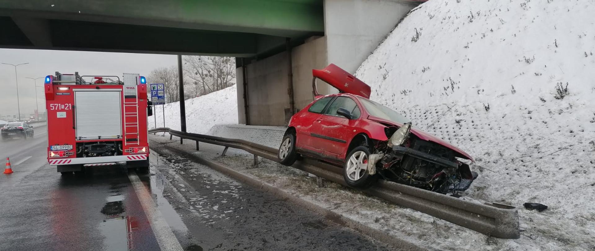 Na zdjęciu znajduje się samochód pożarniczy stojący na prawym pasie drogi po prawej stronie na barierce znajduje się czerwony samochód osobowy. Zdjęcie wykonane zimą.